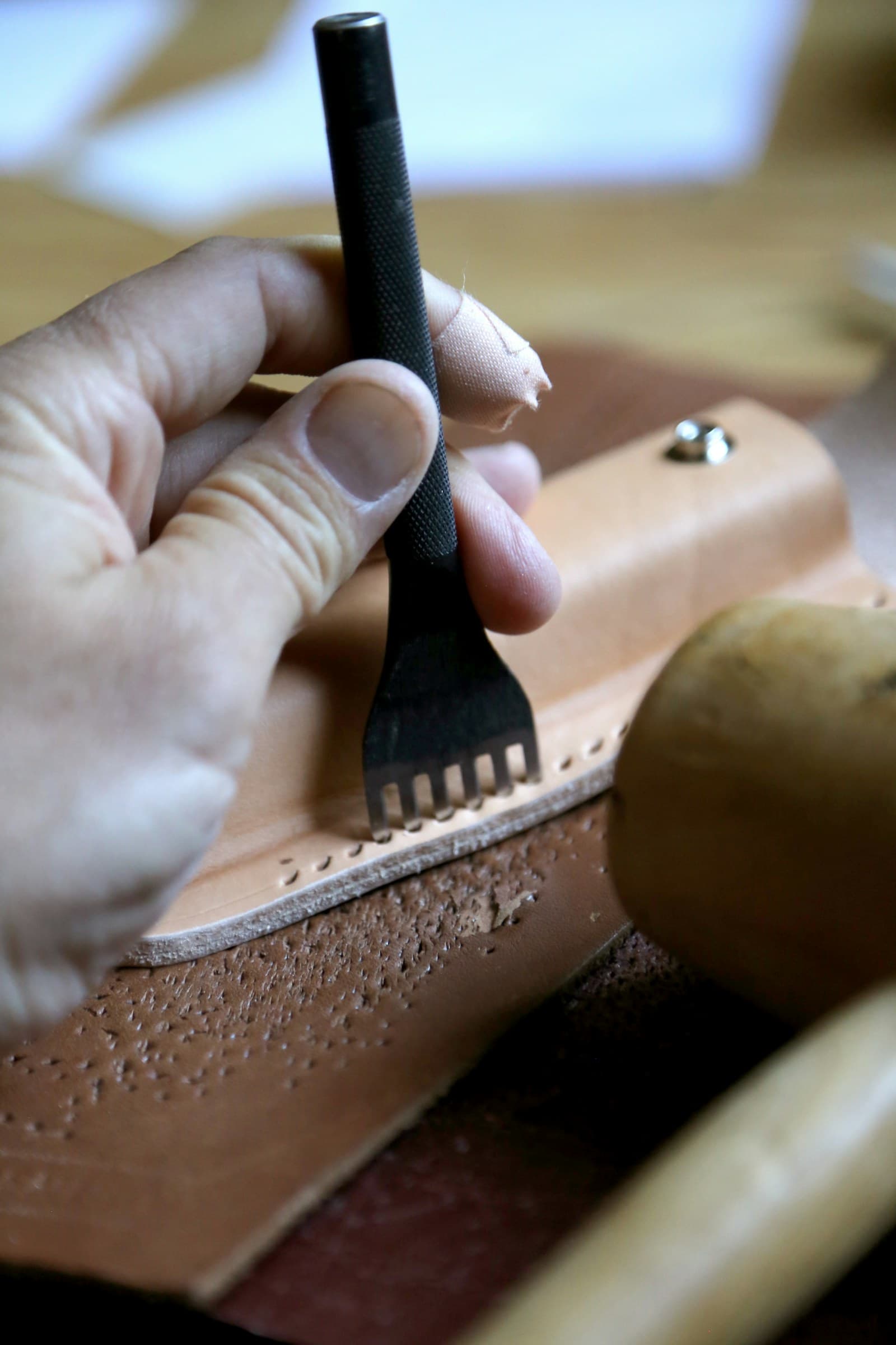 Hands pressing a pricking iron into a pale undyed vegetable-tanned panel, the natural crust colour showing