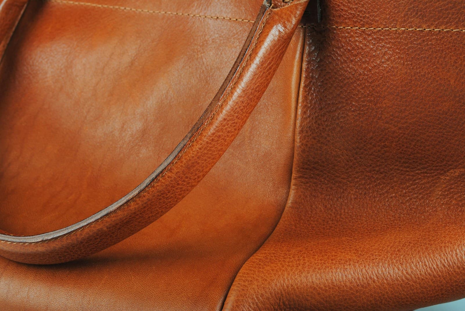 Cognac full-grain hide falling into soft folds around a stitched handle, the pebbled grain lifting where the light rakes across it